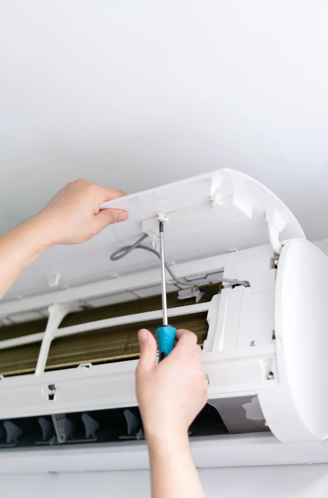 Ductless repair Hands using a screwdriver to repair or install the cover of a wall-mounted air conditioner unit, with exposed internal components visible.